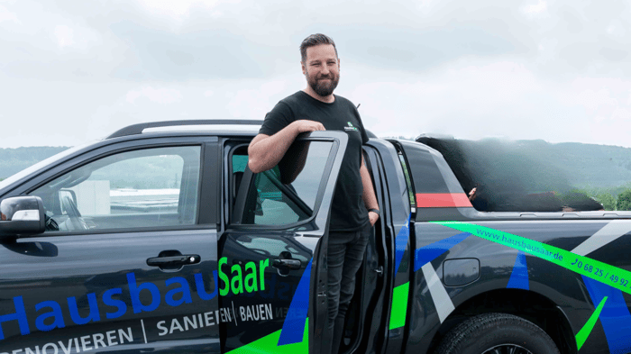 Mann in schwarzem T-Shirt lehnt an offenstehender Tür eines schwarzen Pickup-Trucks mit "Hausbau Saar" Logo.