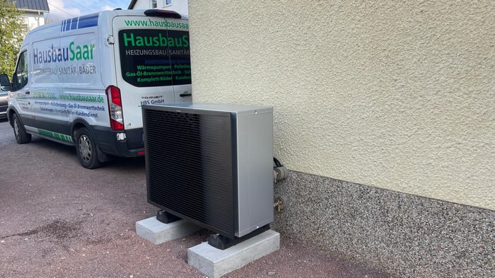 Dunkle Wärmepumpe auf Sockeln an einer Hauswand, daneben ein weißer HausbauSaar-Transporter
